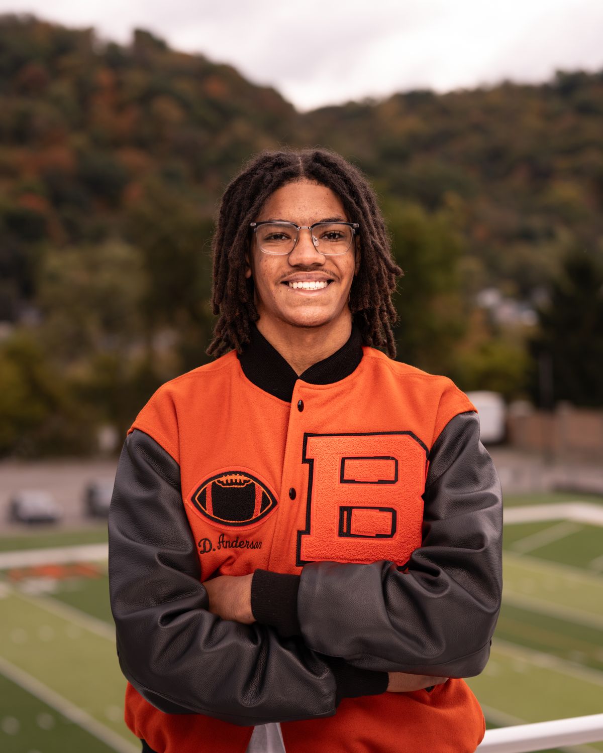 Smiling senior portrait in a letterman jacket by the football field, with autumn hills behind