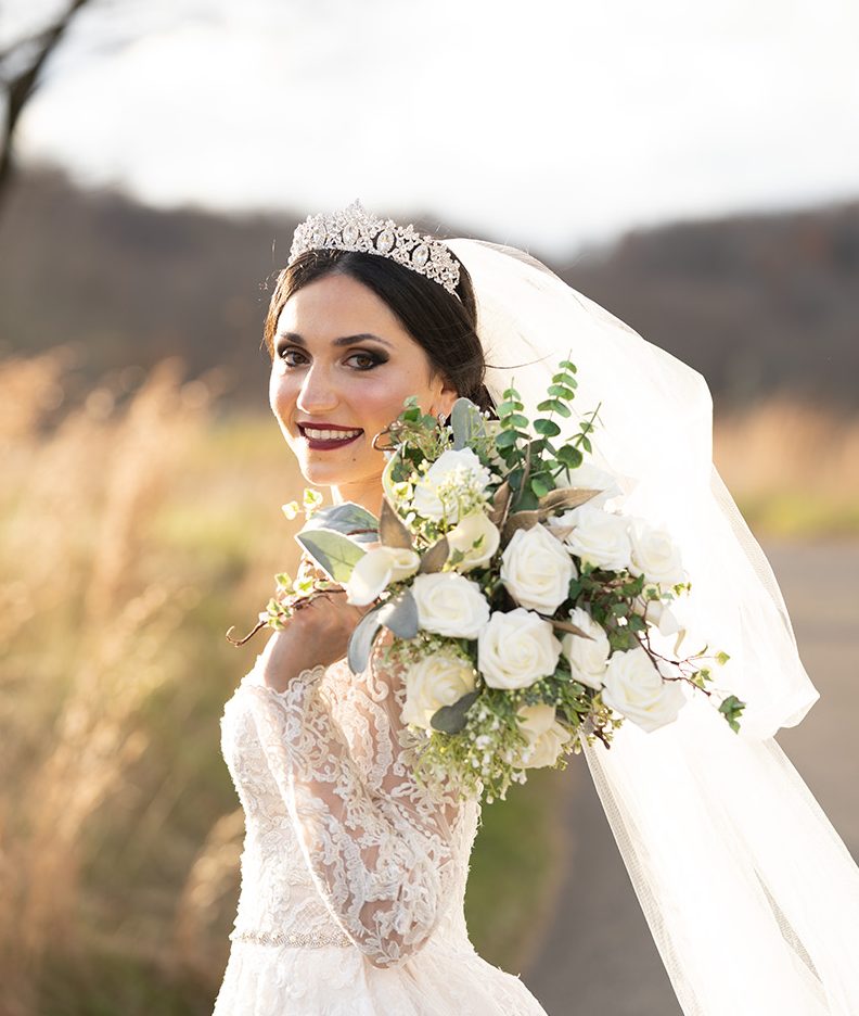 Bride in a lace gown and veil holding a white bouquet, smiling outdoors