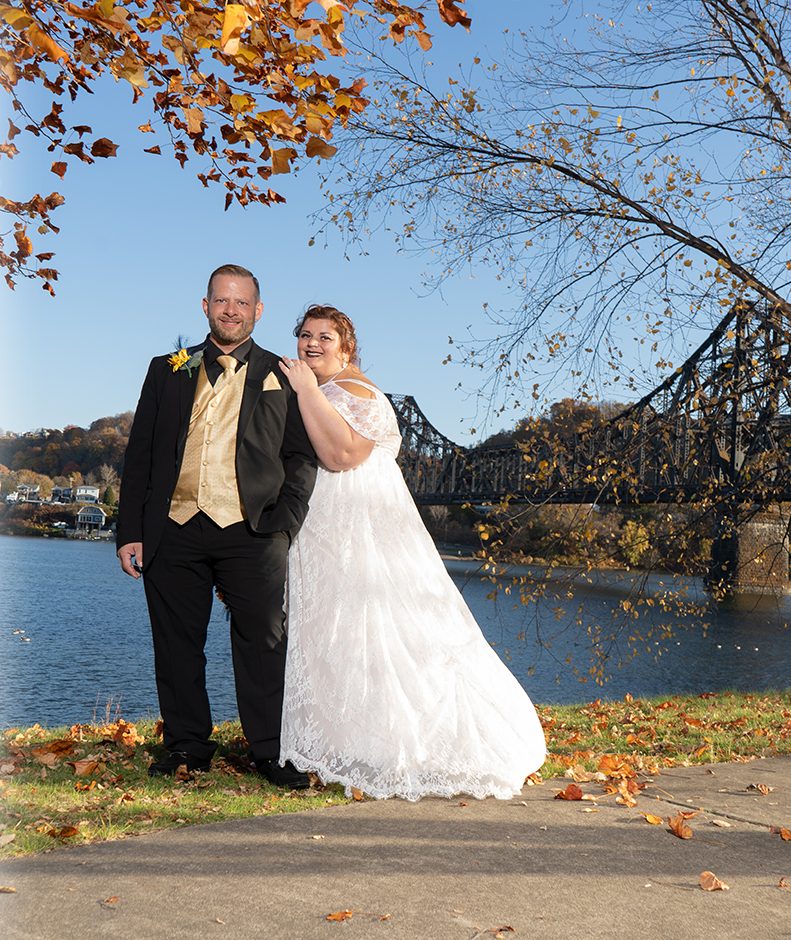 Couple on a riverside path in autumn with a steel bridge behind them, Pittsburgh wedding photography